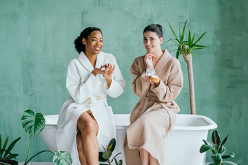 Cheerful pretty multiracial young ladies friends wear silk robes having fun hold glasses with champagne sit on bed in hotel, happy diverse girls bride and bridesmaid celebrate bachelorette spa party