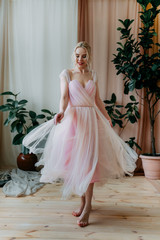 Fototapeta premium A beautiful happy cheerful attractive tender blonde girl in a lush light soft pink spring dress poses twirls and dances for the camera against a background of greenery