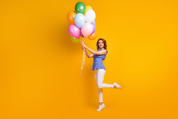 Fototapeta na wymiar Full length profile photo of pretty lady hold many colorful air balloons visit event party wear polka-dot blue shirt white pants gumshoes isolated yellow color background