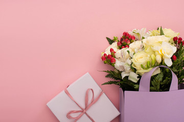 top view of bouquet of flowers in violet paper bag near gift box on pink background
