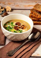 Hot tasty mushroom cream soup decorated with green onion and fried mushrooms in a white bowl on a brown kitchen towel with bread and fresh champignon background