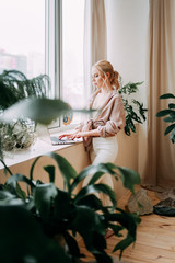 A thoughtful serious girl blogger freelancer works at home on a laptop, writes ideas in her notebook diary, and organizes in a bright spacious office with a view of the city decorated with greenery