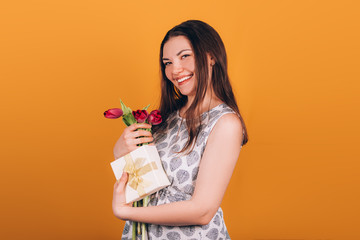 Young beautiful brown-haired girl holding a gift and tulips in hands on an orange background, happy mother's day