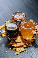 three glasses of expensive craft beer, classic and unfiltered and dark in a glass on the table with a snack of peanut and pistachio chips and nachos