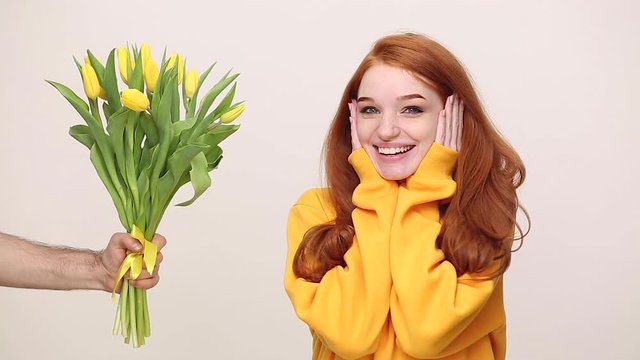 Pretty happy charming ginger redhead young girl in yellow streetwear hoodie posing isolated on white background in studio. People holiday lifestyle concept. Man gives bouquet of spring flowers tulips