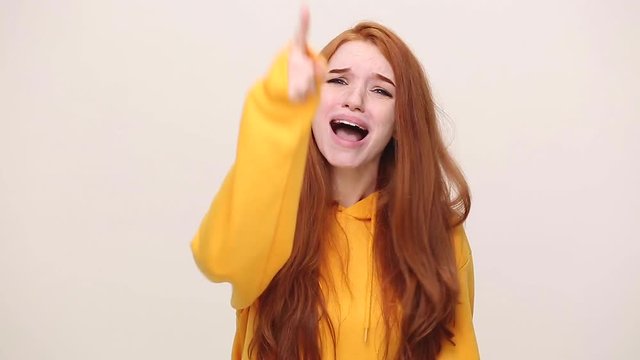 Angry mad ginger redhead curly young girl in yellow streetwear hoodie isolated on white background in studio. People emotions lifestyle concept. Look at camera screaming scolding protest waving hands