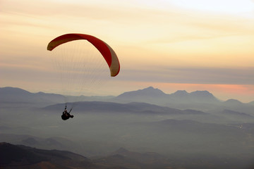 Paraglider flying at sunset in valley with mist and mountains in the background.