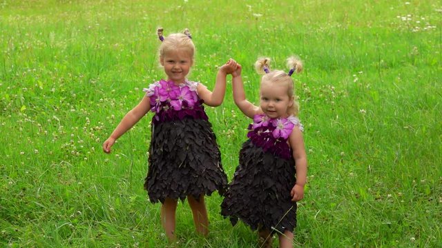 Two blond little sisters in a purple fantasy dresses made of leaves and flowers are holding their hands and dancing on the meadow