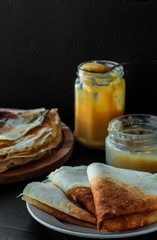 Tasty Thin Pancakes on the white plate with Orange Curd Jar on dark background and a teaspoon of curd and a jar of honey on the dark table vertical orientation
