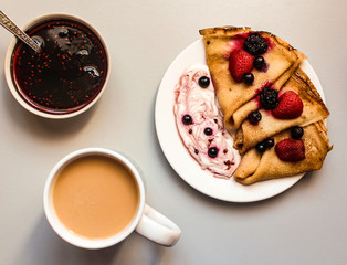 Tasty thin crepes served with sour cream, black currant and fresh strawberries on the table with a cup of tea and strawberry jam horizontal orientation