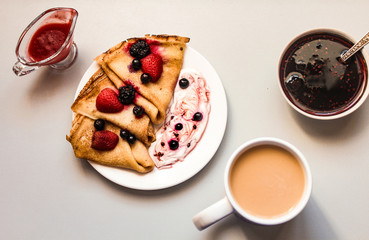 Tasty thin crepes served with sour cream, black currant and fresh strawberries on the table with a cup of tea and strawberry jam and strawberry sauce horizontal orientation