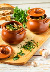 Clay pots with baked potatoes and chicken meat with pumpkin and choped greens on a wooden plank. Garlic and bread on the table
