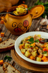 Clay pot with baked potatoes and chicken meat with pumpkin and choped greens. Garlic, pumpkin, bread and greens on the table. Served in a white plate. Vertical orientation