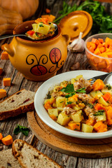 Clay pot with baked potatoes and chicken meat with pumpkin and choped greens. Garlic, pumpkin, bread and greens on the table. Served in a white plate. Vertical orientation