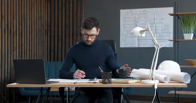 Bearded hipster`s Engineer is drinking Coffee to go on Working Place. Handsome Architect is working in Loft Office, drinking Coffee, using professional Architecture's Tools. Stylish Designer.