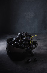 a cluster of juicy black grapes lies in a dark gray bowl on a dark gray table