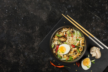 Noodle bowl with mushrooms, egg, tofu cheese, chopsticks and ingredients on a dark background. Asian food.