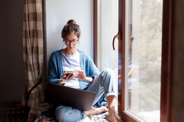 Young woman working from home office. Freelancer using laptop, phone and the Internet. Workplace in living room on windowsill. Concept of female business, career, shopping online. Lifestyle moment. - obrazy, fototapety, plakaty