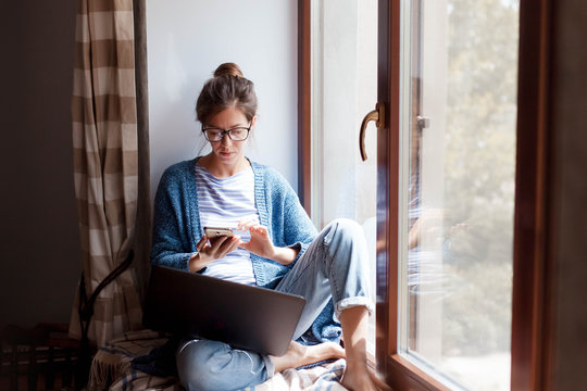 Young woman working from home office. Freelancer using laptop, phone and the Internet. Workplace in living room on windowsill. Concept of female business, career, shopping online. Lifestyle moment.