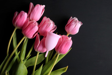 Menstrual cup and pink tulips on a black background. Menstruation, means of protection. Women's health, lifestyle.