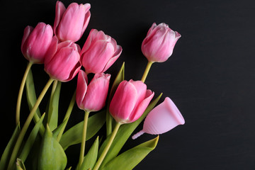 Menstrual cup and pink tulips on a black background. Menstruation, means of protection. Women's health, lifestyle.