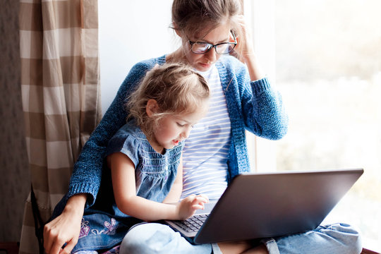Working mom works from home office with kid. Mother and daughter read news. Woman and cute child sitting on window sill. Freelancer workplace with laptop and the Internet. Female business, career.