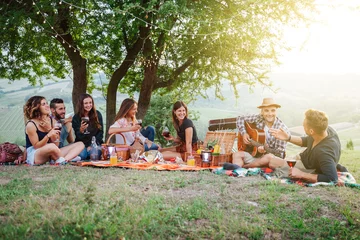 Foto op Canvas Picnic in the countryside - Group of young friends at sunset on spring day are sitting on the ground in a park near trees - They drinking red wine and eating grilled meat with barbecue and play guitar © loreanto