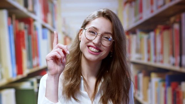 Successful young smart woman staying in the library thinking coming up with useful idea smiling finger pointing posing near shelves with books.