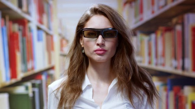 Young brunette woman working in the library wearing black virtual reality glasses using technology in modern education. Background of bookshelves.