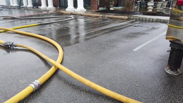 A tangle off fully inflated yellow fire hoses on a wet parking lot fire   scene. There is water flowing into the lot from the building where thew fire   is being fought