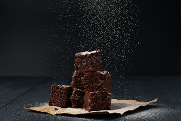 Stack of a brownie pieces on a grey table with dark background with sugar powder snow.