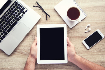 Male hands holding tablet computer with laptop, smartphone and cup of tea on table