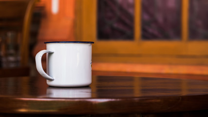 Vintage enamel cup on a classic teak table against simple window background in blur. Antique rustic coffee cup on a brown, smooth wooden surface.
