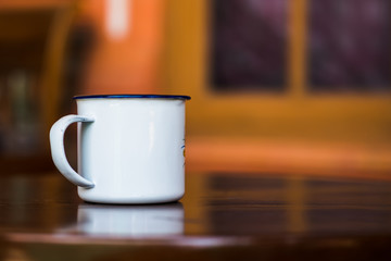 Vintage enamel mug on a classic teak table against simple window background in blur. Antique rustic coffee cup on a brown, smooth wooden surface.