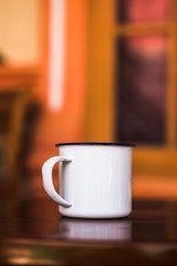 Enamel mug on wood table against blur background of orange window. Antique rustic coffee cup on a brown, smooth wooden surface, suitable for vertical designs.