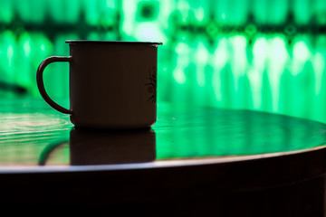 Silhouette of enameled cup on wood table against green fence blur background. Antique rustic coffee cup on a brown, smooth wooden surface.