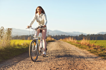 Young woman rides bicycle on dusty road towards camera, afternoon sun shines to fields and forest in background