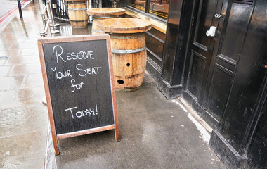 Black Chalkboard with "Reserve your seat for Today" text in front of restaurant or pub black door. Wooden barrel tables standing on wet concrete at background