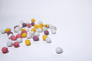 Small sweet candies in the shape of Easter eggs lie randomly on a white background. Different colors - white, yellow, purple, horizontal view
