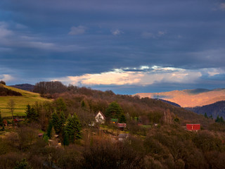 Afternoon sun burns on tops of hills in the distance through a hole in dense dark clouds in the sky