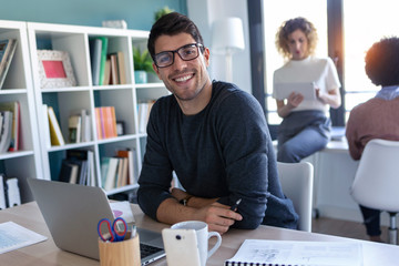 Handsome young entrepreneur looking at camera while working with laptop in the office.