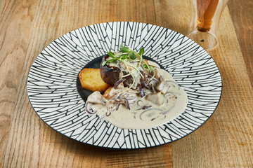 Large portion of delicious beef steak with creamy mushroom sauce and fried potatoes in a ceramic plate on a wooden background.