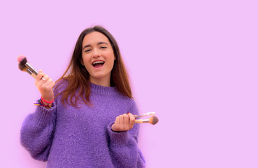 Mujer joven sonriente con brochas de maquillaje en la mano sobre un fondo lila