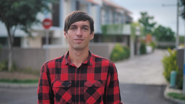 Portrait of handsome caucasian man in red plaid shirt standing in front of the camera and smiling at suburb of the city on the background of houses