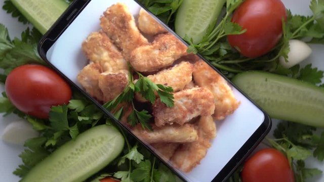 Plate with salad and cellphone with crispy fried cod fish fillet on screen. Food blog concept. Top view of mobile phone with food footage on screen rotating.