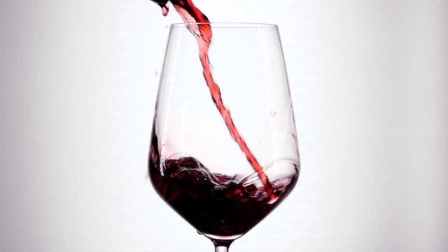Slow motion of pouring red wine from bottle into goblet. Close-up of red wine forms beautiful wave in glass. Wine pouring in glass at white background.