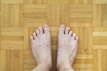 Caucasian male bare feet with crooked toe finger standing on wooden tiles floor top view body hair