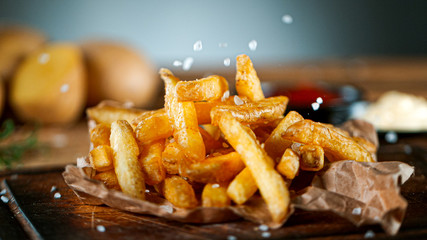 Freeze Motion Shot of Falling Fresh French Fries on Wooden Table and Adding Salt - obrazy, fototapety, plakaty