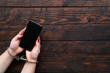 Woman hands in handcuffs hold a modern smartphone on a wooden background. The concept of internet and gadget dependency.