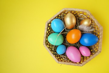Fototapeta na wymiar Golden, pink, blue, green, orange eggs in the wicker basket on the yellow background. Copy space. Place for text and design. Happy Easter. Top view. Flat layout.